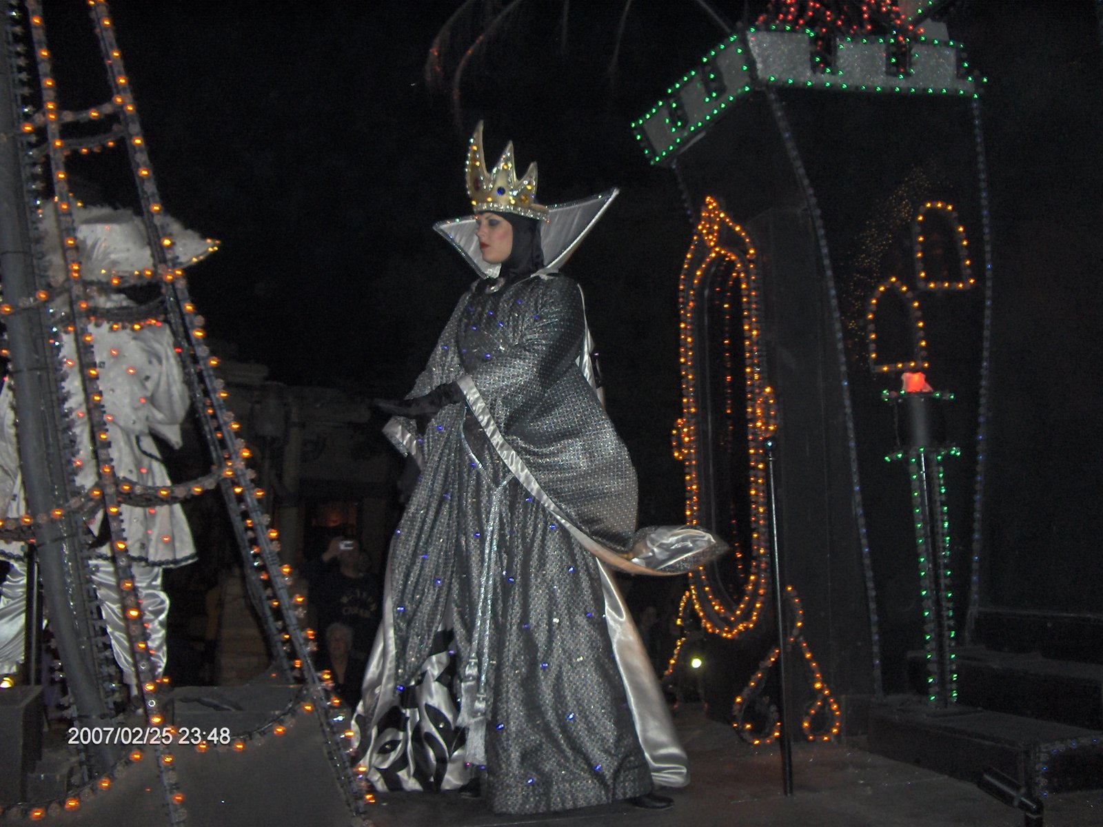 Personagem da Rainha Má em vestido cinza e coroa dourada caminhando no SpectroMagic com colunas de luzes ao fundo