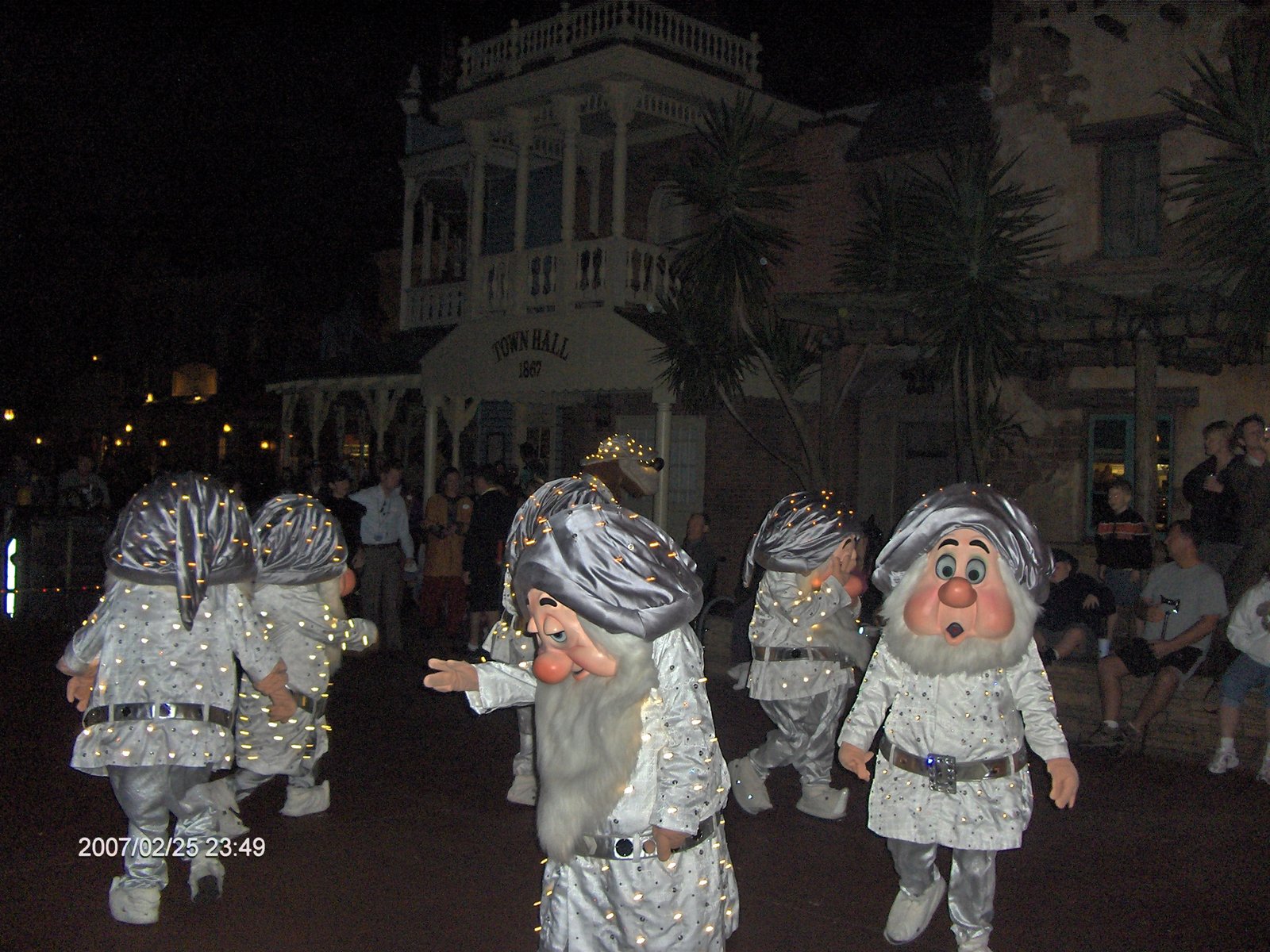 Personagens dos sete anões em roupas prateadas iluminadas caminhando na Main Street em frente ao Town Hall do Magic Kingdom