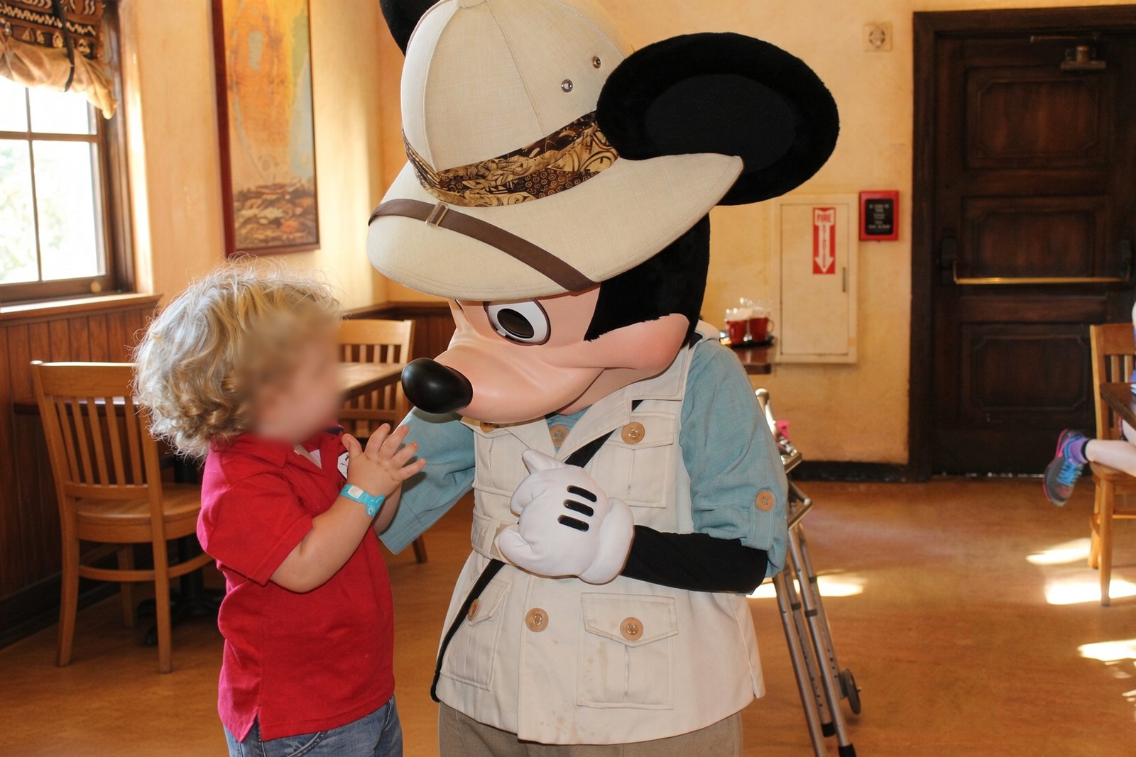 Mickey em roupa de safári cumprimenta uma criança com rosto desfocado no Tusker House